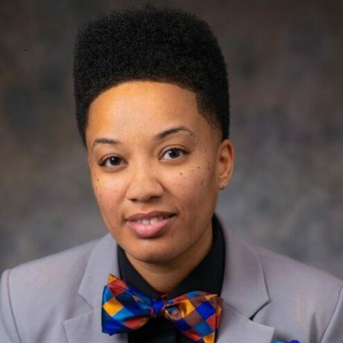 A person with short curly hair in a suit and colorful bow tie smiles softly against a neutral background, conveying a confident and approachable tone. Michelle Strange Director of Bellevue College Restorative Practices