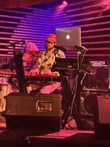 The image captures a moment from a live performance featuring The Outta Towners keyboardist, Jacob Martin. He is seated behind a Nord Stage 3 keyboard, deeply focused on playing, with a laptop set up in front of him. Jacob is wearing a colorful, tropical-print shirt and a red cap that reads “CONSPIRACY THEORIST.” The stage is lit with purples and reds, adding to the lively atmosphere of the performance. In the background, the drummer is partially visible, adding depth to the scene. The setting reflects the intimate, energetic vibe of the venue, with Jacob’s keyboard playing adding layers of warmth and complexity to the band’s sound.