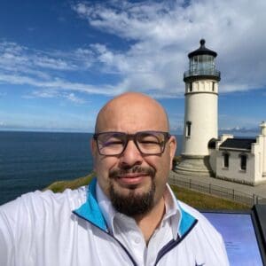 In the image, Richie Martinez is outdoors with a lighthouse in the background on a clear, partly cloudy day. He is smiling, wearing black-rimmed glasses and a white shirt with a blue-collared zipper jacket layered over it. His head is shaved, and he has a well-kept goatee. The lighthouse, standing tall to his right, suggests a coastal location with the sea visible behind him. The atmosphere is calm and scenic, with a sense of nature and open space.