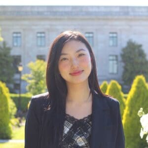 Monyu Yu, shown in the attached image, is a Bellevue College student and regional coordinator for WA-SEN (Washington Student Engagement Network). With a background in International Business Management, she brings a blend of cultural awareness and a passion for creative expression. The image captures her standing outside in a formal environment, wearing a plaid top underneath a blazer. Her hair is long, and her facial expression is serene and confident, reflecting her academic achievements and active involvement in various student organizations at Bellevue College, including the Associated Student Government and Makerspace Club.