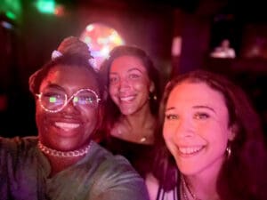The image shows three people smiling at the camera in a warmly lit venue. To the left is Jalisa, wearing large, reflective glasses. She has a bright smile, illuminated by the colorful lighting in the background. In the middle is Keely, radiating a joyful, carefree energy with her wide smile and casual, relaxed appearance. To the right is Morgan Paris Lanza, smiling confidently. Morgan is a talented musician known for her contributions to the local music scene. The lighting gives the scene a vibrant, intimate feel, capturing the camaraderie and fun of the evening. 91.3 KBCS Jalisa Bass
