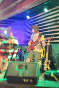 The image captures The Outta Towners bassist, Matthew Crissey, performing live on stage. Matthew is seen playing his bass guitar, wearing a casual outfit of a t-shirt, shorts, and a bucket hat, giving off a relaxed and approachable vibe. He stands slightly turned toward the band, deeply focused on his instrument. In the background, you can see other band members and equipment, including a laptop on a stand and additional instruments. The stage lighting is a mix of vibrant purples and greens, adding a dynamic energy to the scene and reflecting the laid-back yet powerful atmosphere of the performance.
