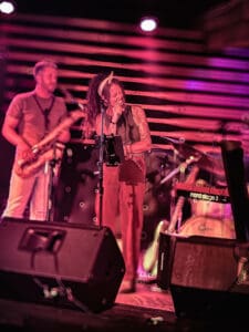 The image captures a live performance by The Outta Towners on stage. In the foreground is vocalist Savanna Woods, energetically singing into the microphone. She wears a headband and has long dreads, with her expressive gestures matching the intensity of her voice. Next to her is the saxophonist, playing alongside the band, bathed in stage lighting. Behind them, you can see part of the drum set and a Nord Stage 3 keyboard, as well as other instruments that contribute to the dynamic atmosphere. The blurred, vibrant lighting gives the scene a lively, electric energy, perfectly encapsulating the raw passion of their performance.