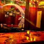 The image shows a cozy, dimly lit area of a tavern, likely a booth seating section with a whimsical mural of a crescent moon with a face looking toward a small table. The wooden walls are adorned with framed photos and decorations, including a mounted fish. The lighting is warm, casting an inviting glow from small candles on the tables and bar. A door marked “HEAD” can be seen in the background. The overall ambiance reflects a nostalgic, quirky vibe with a blend of retro decor, making it an intimate space for gatherings.