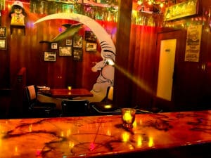 The image shows a cozy, dimly lit area of a tavern, likely a booth seating section with a whimsical mural of a crescent moon with a face looking toward a small table. The wooden walls are adorned with framed photos and decorations, including a mounted fish. The lighting is warm, casting an inviting glow from small candles on the tables and bar. A door marked “HEAD” can be seen in the background. The overall ambiance reflects a nostalgic, quirky vibe with a blend of retro decor, making it an intimate space for gatherings. 91.3 KBCS Jalisa Bass