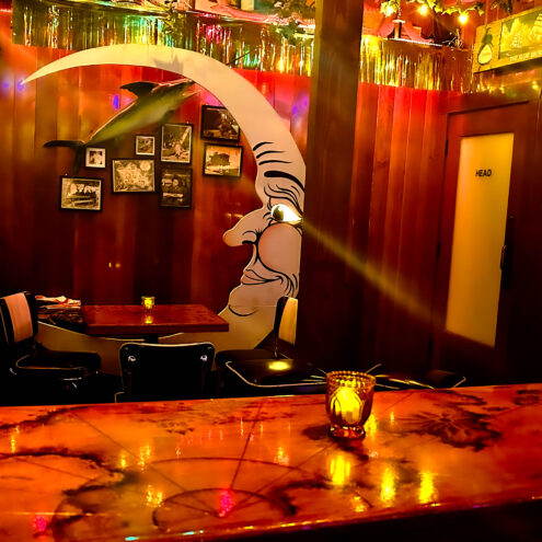 The image shows a cozy, dimly lit area of a tavern, likely a booth seating section with a whimsical mural of a crescent moon with a face looking toward a small table. The wooden walls are adorned with framed photos and decorations, including a mounted fish. The lighting is warm, casting an inviting glow from small candles on the tables and bar. A door marked “HEAD” can be seen in the background. The overall ambiance reflects a nostalgic, quirky vibe with a blend of retro decor, making it an intimate space for gatherings.