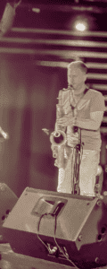 The image features a saxophonist performing live on stage in a dimly lit venue. The musician, blurred slightly in motion, stands with a saxophone in hand, positioned near a microphone stand. He is in front of a striped black and white background with soft, ambient lighting. Two large stage speakers are visible in the foreground, emphasizing the live performance setting. The image conveys the energy of live music in an intimate, moody atmosphere.