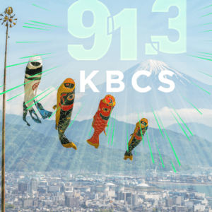 The image features a scenic view with the KBCS 91.3 FM logo overlaid. In the background, Mount Fuji stands tall with snow-capped peaks, set against a clear blue sky. Below the mountain, colorful koinobori (carp-shaped windsocks) are flying in the wind. These are traditional Japanese decorations typically flown to celebrate Children’s Day during Golden Week. The foreground showcases a city landscape with a mixture of buildings and natural greenery.

This visual highlights both Japanese culture and the iconic nature of Mount Fuji, suggesting a celebration or event connected to Japanese traditions. Clicking the image takes the viewer to the Bellevue College Japan Week Flyer for more information.