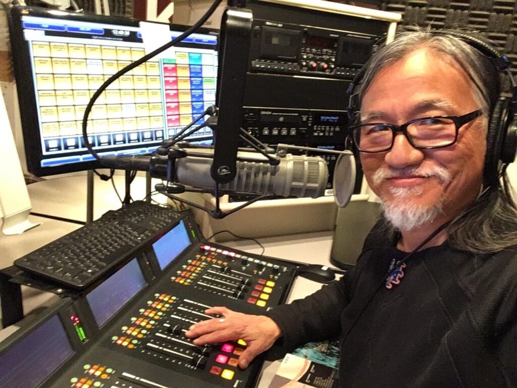 A photo of John Pai in the mixing room at KBCS. He is an asian man with long grey hair and a short goatee smizing while looking into the camera. He is using professional audio equipment, including a microphone, headphones, and a mixing board.