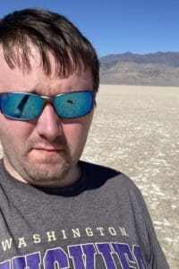 Male with sunglasses on against a backdrop of flat ground and mountains in the distance.