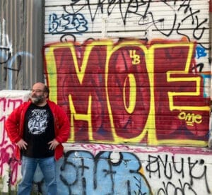 A photograph of a man with light skin wearing a graphic t-shirt, a red jacket, and jeans. He is standing in front of a wall of graffiti which has, "MOE", spray painted on in large all caps text with yellow outline and a red fill.
