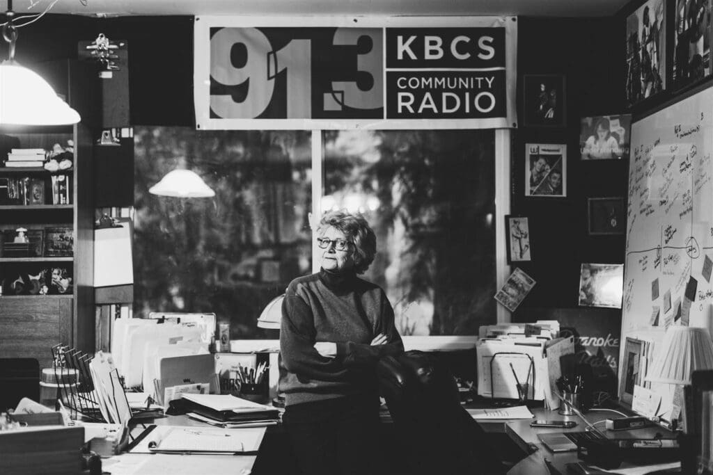 Black and white image of Auntmama in her KBCS office. She is a light skinned woman with glasses positioned in the center of the photograph in front of the window in a confident pose with her arms crossed and head turned slightly to the left. The office is cluttered with papers, images, a bookshelf on the left, a whiteboard on the right, and a large KBCS banner above the window.