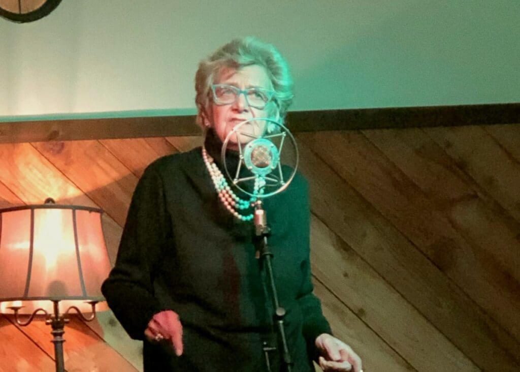 Photograph of Auntmama performing live in front a microphone. She is a light skinned woman with gray hair wearing glasses, gold pearl earrings, a black turtleneck, and three white pearl necklaces. Her expression is pensive and contemplative with her eyes looking up and fingers pointed down.