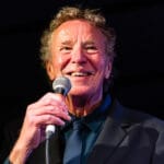 John Gilbreath, the host of the Caravan on KBCS, in a dark suit and blue shirt holds a microphone, smiling warmly against a dark background, conveying a joyful and lively mood.