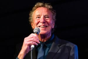 Man in a dark suit and blue shirt holds a microphone, smiling warmly against a dark background, conveying a joyful and lively mood.