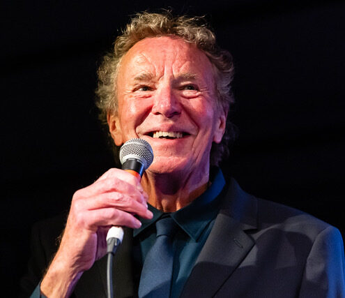 John Gilbreath, the host of the Caravan on KBCS, in a dark suit and blue shirt holds a microphone, smiling warmly against a dark background, conveying a joyful and lively mood.