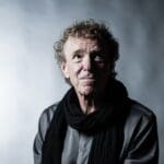 Man with fair skin, brown blond hair wearing grey shirt and black scarf in front of grey wall