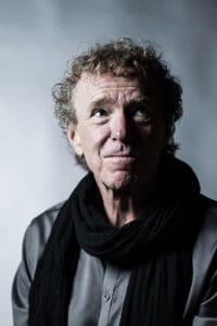 Man with fair skin, brown blond hair wearing grey shirt and black scarf in front of grey wall