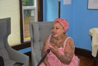 A person with tattoos, wearing a red and white dress and headscarf, sits on a gray chair, speaking into a microphone in a blue-walled room.