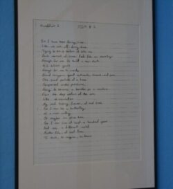 A framed handwritten poem on lined paper against a blue wall. The text is neatly written in black ink, conveying a reflective and creative tone.