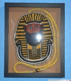 Framed beaded artwork of an ancient Egyptian pharaoh's mask in gold, black, and white, with a yellow rope accent, set against a blue wall.