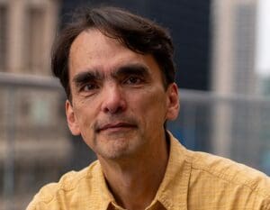 Portrait of a middle-aged man in a yellow shirt, with a neutral expression. He is outdoors with blurred city buildings in the background.