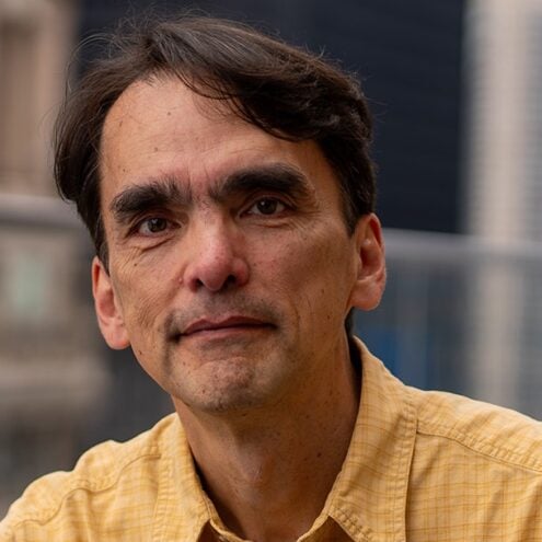 Portrait of a middle-aged man in a yellow shirt, with a neutral expression. He is outdoors with blurred city buildings in the background.
