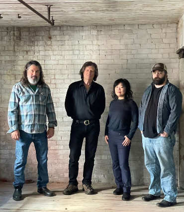Four people stand in a dimly lit room with white brick walls and wooden floors. They appear serious, dressed in casual and dark clothing.