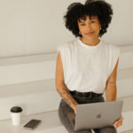 A person with curly hair smiles while seated on white stairs. They're using a laptop, wearing a white sleeveless top and jeans, with a phone and coffee cup beside them.