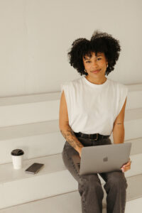 A person with curly hair smiles while seated on white stairs. They're using a laptop, wearing a white sleeveless top and jeans, with a phone and coffee cup beside them.