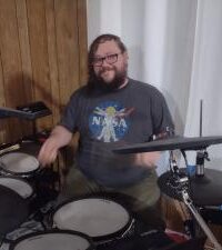 John Weir, bearded guy with glasses in a grey NASA Tshirt sitting behind drum kit