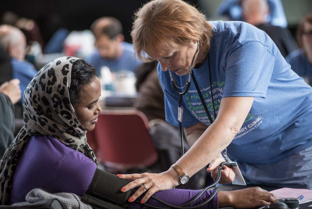 A healthcare worker in a blue shirt takes the blood pressure of a woman in a purple top and patterned headscarf. The setting is a busy clinic.