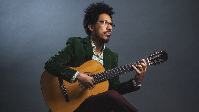 Man with glasses and an afro haircut sits thoughtfully playing a classical guitar. He's wearing a green velvet jacket. The background is a muted gray.