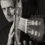 A man gazes intensely while holding a guitar upright. The black-and-white tones create a dramatic, introspective mood, focusing on musical passion.