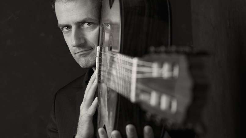 A man gazes intensely while holding a guitar upright. The black-and-white tones create a dramatic, introspective mood, focusing on musical passion.