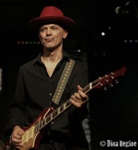 A musician in a red hat plays an electric guitar on stage. He wears a black shirt, focused expression, dimly lit background, conveying a moody, intimate vibe.