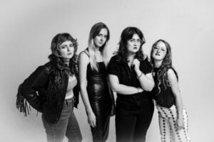 A black-and-white photo of four women standing confidently and posing. They wear eclectic, edgy outfits with leather and fringe accents. The mood is bold and expressive.