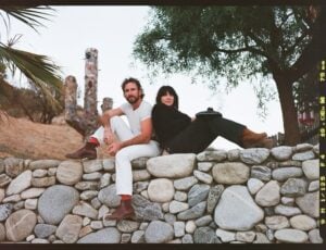 A man in white and a woman in black sit comfortably on a stone wall, surrounded by a desert landscape, palm trees, and a tree, conveying a relaxed, bohemian vibe.