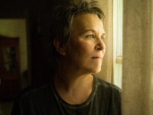 A person with short hair gazes thoughtfully out a sunlit window, soft light highlighting their face. The scene conveys a calm, reflective mood.