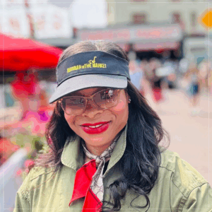 Madinah Slaise in visor cap with Madinah in Market logo on it. Long black hair, sunglasses, red lips in light green top and scarf with red end.
