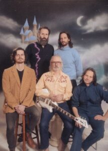 Formal group portrait of Rose City Band featuring five men posed in front of a painted backdrop showing a moonlit castle scene with one member seated holding a white electric guitar.