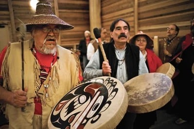 Two Indigenous men in traditional regalia play large hand drums during a ceremonial gathering inside a wooden longhouse style building with other community members visible in the background