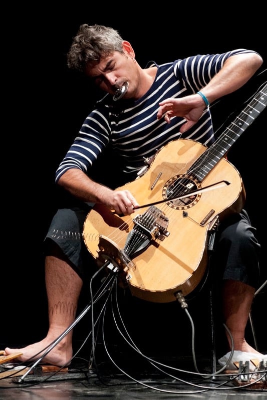 Paolo Angeli performing live on stage playing his heavily modified prepared Sardinian guitar with a bow wearing a striped shirt and white pants surrounded by cables and electronic equipment on the floor against a large black backdrop