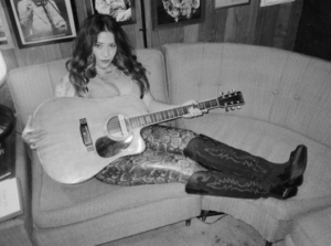 Black and white press photo of Hannah Juanita a woman with long curly dark hair reclining on a couch holding an acoustic guitar and wearing snakeskin print pants and tall cowboy boots in a vintage decorated living room