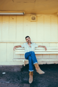 Press photo of Dylan Earl a man with a mustache sitting on a white bench under a yellow painted overhang wearing a light patterned button up shirt blue jeans and tan boots with one leg crossed over the other