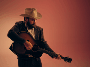 Press photo of Mose Wilson a bearded man wearing a tan cowboy hat and dark pinstripe suit holding an acoustic guitar with both hands extended to the side against a warm gradient orange and pink background