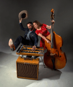 Press photo of Poi Rogers duo Gerard Egan reclining backward on a steel guitar wearing a cowboy hat in motion while Carolyn Sills in a red dress plays an upright double bass beside him against a plain grey studio background