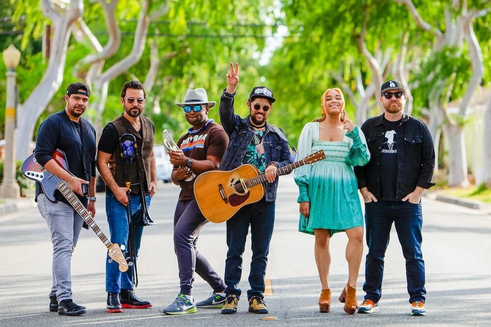 Dustbowl Revival band press photo showing six members standing in the middle of a tree lined street in Los Angeles with two guitarists on the left one member holding a trumpet at center a frontwoman in a teal dress and a guitarist on the right all posing casually in bright summer light