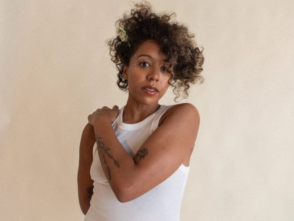 Press photo of Lizzie No a Black woman with short natural curly hair adorned with small white flowers wearing a white sleeveless ribbed top with arms crossed and small crystal face gems photographed against a warm neutral beige background with a direct steady gaze toward the camera