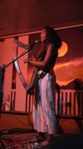 Lizzie No a Black woman with curly hair performing live on stage playing a standing harp and singing into a microphone wearing a black sleeveless top and white wide leg pants bathed in warm orange and red stage lighting against a painted sunset mural backdrop